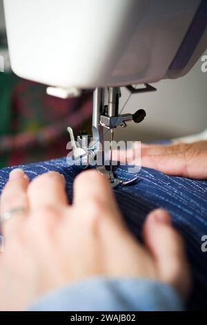 A detailed shot of a sewing machine needle and presser foot on blue denim, with a colorful pin cushion in the background. Stock Photo