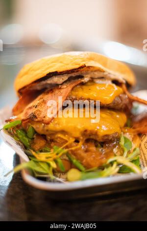 Gourmet beef burger with melted cheddar, crispy bacon, caramelized onions, and fresh greens on a brioche bun, served on a newspaper-lined plate Stock Photo