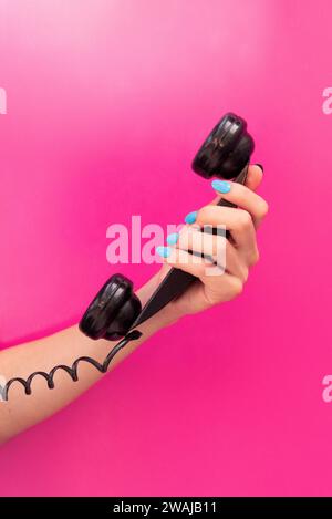 Anonymous woman's hand with blue nail polish holding a black telephone receiver against a pink background Stock Photo