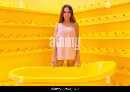 Young woman standing in a yellow bathtub in a coordinated yellow room adorned with rows of rubber ducks looking at camera Stock Photo