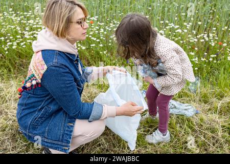 High angle of a blonde woman kneeling and holding a garbage bag with a young unrecognizable girl collecting plastic waste in a wildflower meadow Stock Photo
