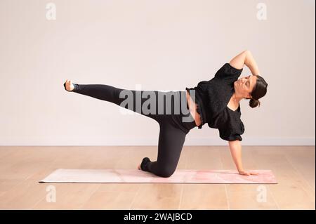Sportive young lady in black activewear stretching arm and leg on mat during yoga workout in spacious room Stock Photo