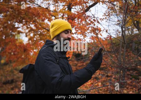 Side view of bearded young male in warm clothes with beanie cap and backpack browsing smartphone while standing by autumn trees in Stockholm City Nati Stock Photo