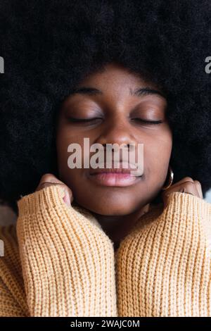 Portrait of cheerful young African American female with Afro hairstyle on a gray background with his hands hidden in his sweater resting on his face w Stock Photo
