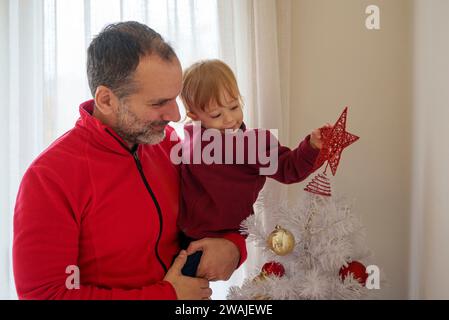 16 month old toddler helping his father set up festive decorations on the Christmas tree Stock Photo