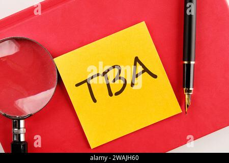 TBA the inscription on the sticker lying on a red notebook next to a magnifying glass and a fountain pen Stock Photo
