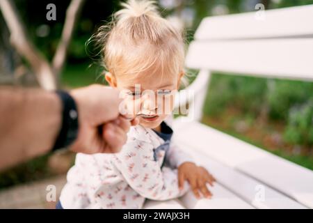 Dad feeds porridge from a spoon to a little girl leaning on a park bench Stock Photo