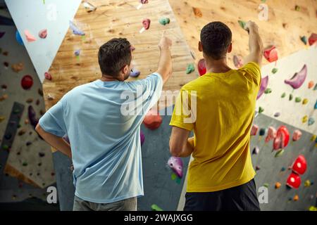 Man standing with instructor, looking and pointing on climbing wall, discussing techniques of climbing, bouldering activity. Stock Photo