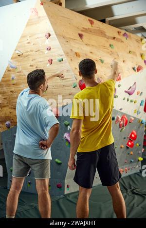 Man standing with instructor, looking and pointing on climbing wall, discussing techniques of climbing, bouldering activity. Stock Photo