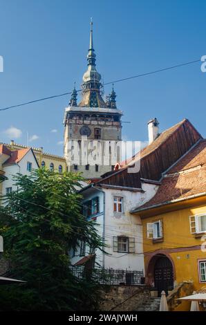 Historical old medieval clock tower surrounded by yellow and white painted houses, buildings with tiled roofs and brick chimneys in Sighisoara Romania Stock Photo