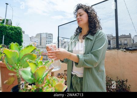 young Latin woman with curlers and green clothes, happy standing with water sprayer wetting plants in terrace garden, taking care of plants by hydrati Stock Photo