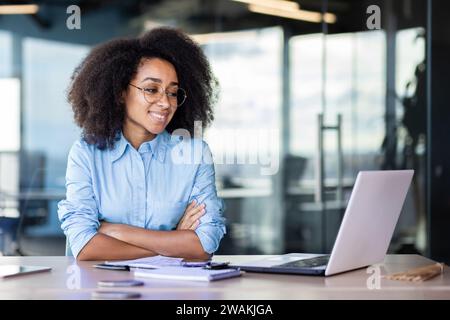 A smiling young African-American woman is sitting in the office at the desk and looking at the laptop screen. Works, studies, talks via video call. Stock Photo