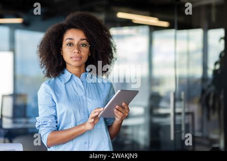 Portrait of an African-American woman standing in the office, holding a tablet in her hands, confidently and seriously looking at the camera. Stock Photo