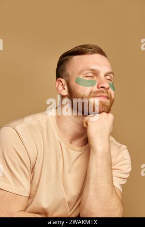 Close up portrait of young attractive man sitting with hydrogel patches under the eyes against swelling and bruising against beige background. Stock Photo