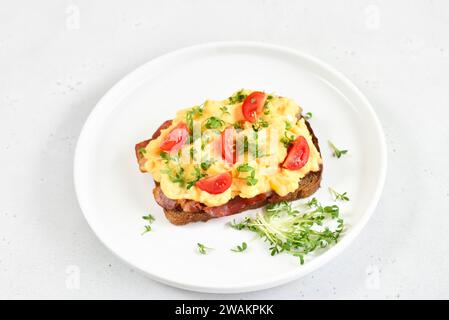 Scrambled eggs with microgreen, bacon and tomato slices on bread, close up Stock Photo