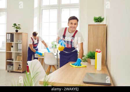 Two happy professional janitors in uniforms cleaning office tables with sanitizer Stock Photo