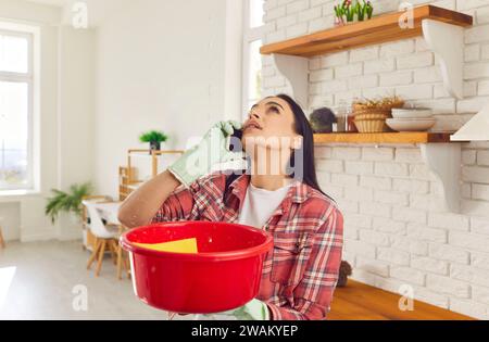 Woman with leaking ceiling at home is collecting water in basin and calling repair service Stock Photo