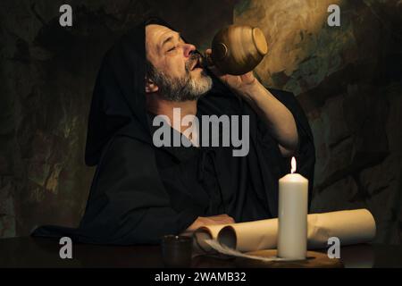 An elderly monk drinking from a clay jug, a dark and smoky stone cell, a candle burning. Stock Photo