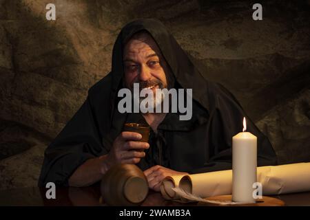 Cheerful monk in cassock drinking wine from a clay goblet sitting at a table, a jug of wine and drinking in a medieval inn. Concept: drunkenness and a Stock Photo