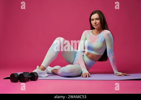 Beautiful long haired sportswoman, wearing activewear, posing in studio. Front view of pretty Caucasian female athlete sitting on floor, smiling to camera after workout. Sport, lifestyle concept. Stock Photo