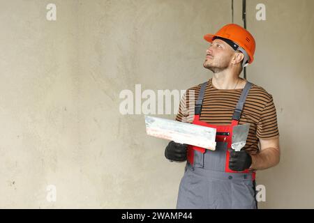 repairman in overalls and protective orange helmet holds two spatulas for plastering concrete wall with putty using. Repair work at apartment and cons Stock Photo