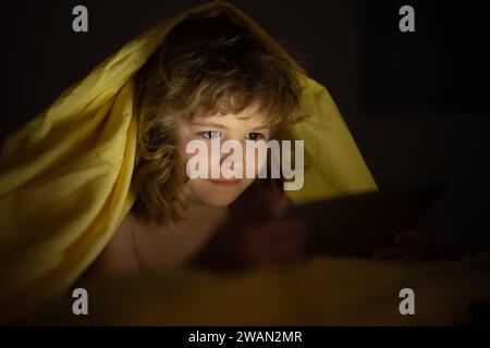 Kid playing on a digital tablet. Kid using tablet for gaming and online learning while lying on the bed, screen light reflex on her face. Child play Stock Photo