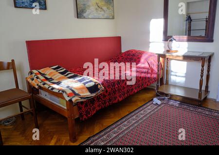 Old cabin type bedroom with small single bed, rustic red bed cover and rug, illuminated by bright sunshine from window. Stock Photo