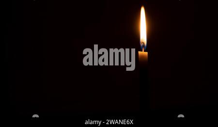 A single burning candle flame or light is glowing on an orange candle on black or dark background on table in church for Christmas, funeral Stock Photo