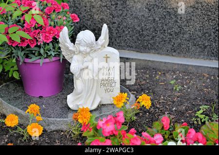 Grave angel with inscription "May an angel protect you" rests on granite slab surrounded by flowers Stock Photo