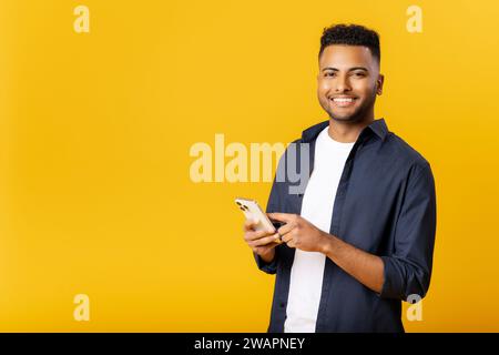 Online communication. Handsome happy indian man using smartphone standing isolated on yellow, arab male student guy scrolling, checking notifications, working on the distance, texting Stock Photo