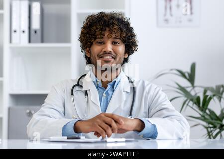 Close-up portrait of doctor inside clinic at workplace, man in white medical coat smiling looking at camera, satisfied indian man with curly hair. Stock Photo