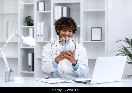 Young successful hispanic doctor using phone, man reading educational material using app on smartphone, working inside clinic medical office, smiling contentedly, sitting with laptop. Stock Photo