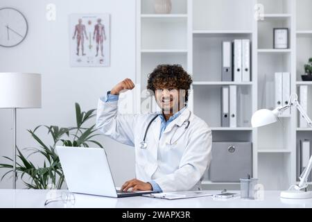 Well done superhero doctor, man smiles looks at camera, holds his hands up, rejoices at the success in the treatment of the patient, works inside the clinic in the medical office, sits at the table. Stock Photo