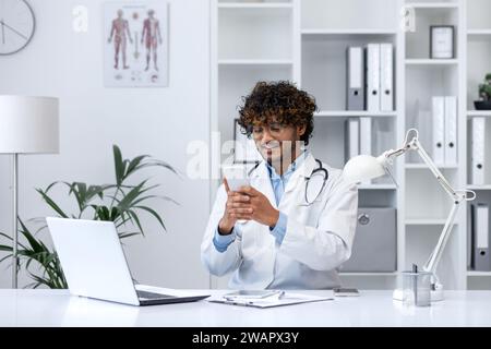 Young successful hispanic doctor using phone, man reading educational material using app on smartphone, working inside clinic medical office, smiling contentedly, sitting with laptop. Stock Photo