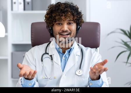 Webcam view, young doctor with headset phone using laptop for video call, doctor cheerfully and friendly consulting patients, smiling and looking at camera, working inside clinic medical room. Stock Photo