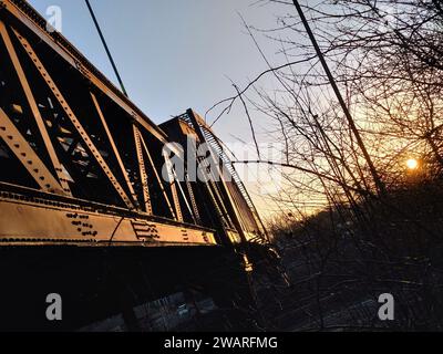 A bridge above a motorway with the sun setting off to the side, tree branches visible in the foreground and the shape of lampposts in the background Stock Photo