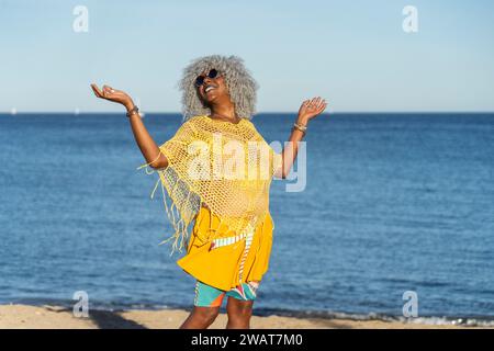 Self-confident, black lady with hoary afro hair and sunglasses smiling while walking on a sunny beach. Concept: pro-aging, vacations Stock Photo