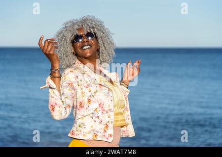Self-confident, black lady with hoary afro hair and sunglasses smiling while walking on a sunny beach. Concept: pro-aging, vacations Stock Photo