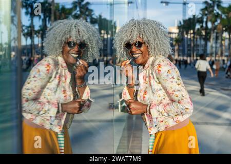 Self-confident, black lady with hoary afro hair and sunglasses putting on lipstick using window as a mirror in sunny city center. Concept: pro-aging, Stock Photo