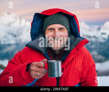 Portrait of smiling at camera high altitude mountaineer dressed red warm dawn jacket holding metal mug of hot tea in with mountains panorama backgroun Stock Photo