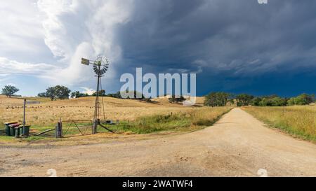 A dark stormfront approaching over a windmill beside a country road and farmland at Yapeen, in Central Victoria, Australia. Stock Photo