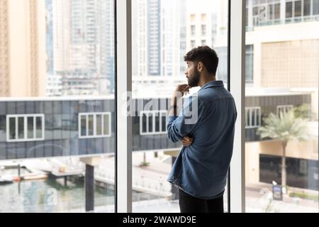 Thoughtful serious handsome young Arab man standing at large window Stock Photo
