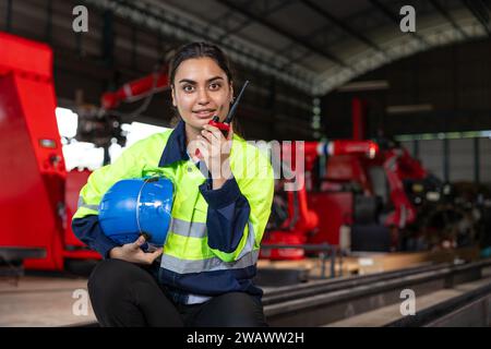 Engineer woman smart female indian worker supervisor with safety suit work in robot metal machinery factory operator radio control. Stock Photo