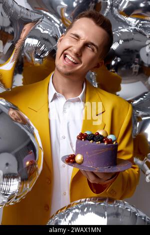 handsome birthday guy in yellow jacket holding cake looking at the camera being lovely on silver helium balloons background. Stock Photo