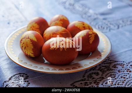 The image shows seven brown eggs with yellow leaf patterns on a white plate with a gold rim, placed on a blue and white lace tablecloth. Stock Photo