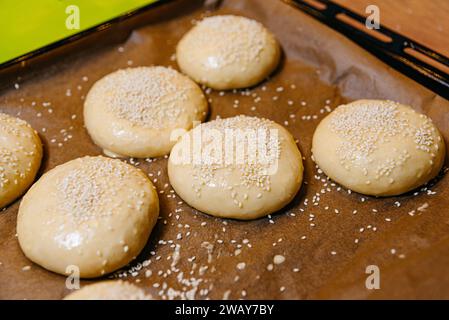 Raw dough for buns topped with sesame seeds ready to be baked on a parchment-lined tray. Stock Photo