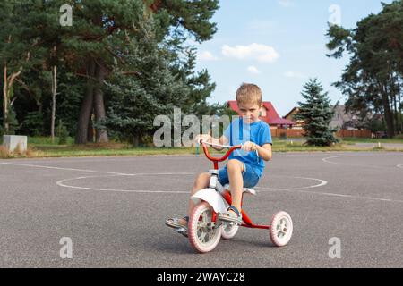 A little boy in blue clothes rides an antique tricycle on an outdoor basketball court. Stock Photo