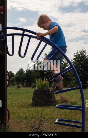 Little boy climbs up the curved ladder on the playground. Child climbs confidently up the ladder against the blue sky background. Stock Photo