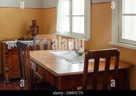 An antique wooden table and three chairs by the window, daylight streaming in to illuminate a baptismal vase and a large antique clock on the table. Stock Photo