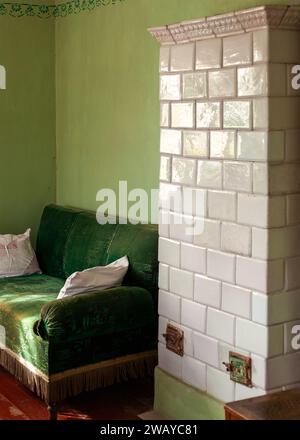 White glazed brick antique wood-burning stove wall and green velour sofa nearby. Vertical photo of a fragment of an ancient interior. Stock Photo